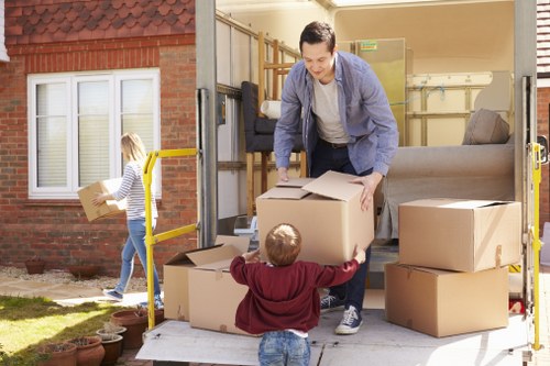 A man wearing casual clothing is seen loading a cardboard box into the back of a moving van parked outside a residential house in West Kensington. The van's rear door is open, revealing additional boxes stacked inside, some on the ground and others being handled by the man. A young child in a red hoodie is assisting by holding a box, while a girl with a striped shirt and jeans carries a smaller box towards the van from the lawn, which features potted plants and a brick wall with a window. The background shows the exterior of a suburban house with brick walls, white-framed windows, and a small garden area. The scene is illuminated by natural daylight, emphasizing the activity of a house removal or moving process in a typical London residential setting, consistent with local removals services in West Kensington. This detailed description aligns with environmental and procedural elements related to house removals, portraying household furniture, cardboard boxes, and outdoor surroundings appropriate for a professional moving company’s website content.