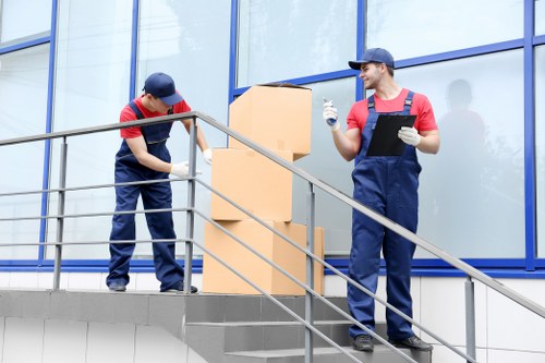 Two professional removal workers dressed in navy blue uniforms and red t-shirts are positioned on an exterior staircase of a modern residential building with large glass windows reflecting the sky. The worker on the left is focused on lifting or securing a large cardboard box, which is placed on top of a smaller box or container, while the worker on the right is smiling, holding a black clipboard or tablet, and wearing white gloves, indicating active communication or planning during the moving process. The staircase has metal railings, and the background suggests a clean, urban residential environment, possibly in West Kensington or nearby in London. The scene captures a moment of careful handling of household items, with attention to safety and efficiency typical of a house removals service in the area. The lighting is natural, with soft daylight illuminating the scene, emphasizing the professional yet approachable nature of the removal team from West Kensington Man with Van.