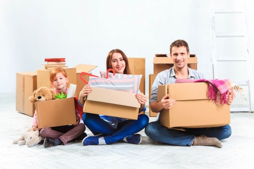 A family of three, consisting of a young girl, a woman, and a man, are seated on the floor of a bright, minimally decorated room with white walls and wooden flooring, surrounded by multiple cardboard boxes in various sizes, some open and filled with household items. The young girl holds a box containing a plush teddy bear and a neatly stacked pile of books. The woman, positioned in the center, is holding a box with clear plastic wrapping and red ribbon, and appears to be smiling. The man on the right is holding a large open cardboard box with a colorful knitted blanket draped over its edge. Behind them, additional boxes are stacked, and a white door or window is partially visible on the right side of the image. The scene suggests they are preparing for a house move or decluttering, with various household belongings ready for transportation or storage. The setting indicates a residential environment, possibly in West Kensington, London, given the context of local house removals services by West Kensington Man with Van.