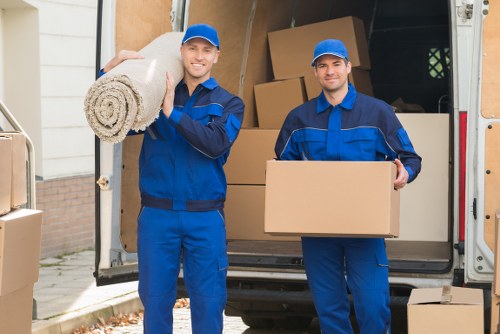 Two professional removals workers dressed in blue uniforms and matching caps are standing outside the back of a white removals van on a residential street near a house with a brick and white exterior. The worker on the left is smiling and carrying a large rolled-up carpet or rug over his shoulder, while the worker on the right is holding a medium-sized cardboard box. Behind them, the van's open rear doors reveal stacked brown cardboard boxes of various sizes, some aligned along the interior sides and others piled in the middle. The scene includes elements typical of house removals, such as the cardboard boxes used for packing personal belongings, and the workers are preparing to load or unload items, consistent with professional relocation services in London or nearby areas. The lighting suggests daytime, with natural light illuminating the scene, contributing to a professional and organized environment suitable for a house move in West Kensington or nearby boroughs.