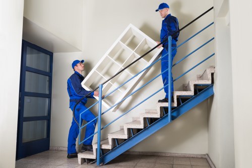Two professional movers from West Kensington Man with Van are carrying a white wooden wardrobe with multiple compartments up a staircase inside a residential building in a quiet area near W14 postcode. The staircase has light wood steps and a blue metal railing, with one mover at the bottom lifting the furniture with both hands, coordinated with the second mover at the top guiding and steadying it. Both movers are dressed in matching blue uniforms and caps, wearing gloves for safety. The interior has neutral-colored walls, a large window with blue frames on the left allowing natural light to illuminate the scene, and gray tiled flooring beneath the window. The environment appears clean and organized, suitable for house removals or estate clearance services, with the focus on safely transporting household items. The scene effectively highlights the careful handling of furniture during a residential move within London’s west Kensington area, emphasizing professionalism in home removals and sustainability practices.