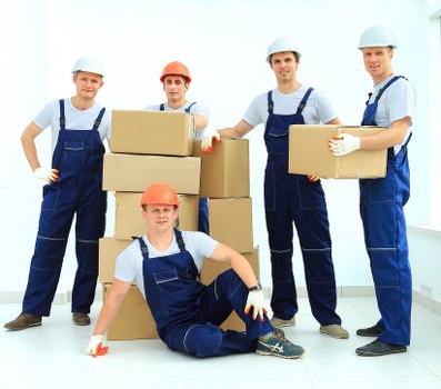 A group of five professional removals workers dressed in blue overalls and safety helmets, with one wearing an orange helmet, posing indoors inside a well-lit, modern residential property. They are surrounded by neatly arranged cardboard boxes, some stacked on top of each other, with one worker kneeling in front and the others standing behind, all smiling at the camera. The boxes vary in size and are used for packing personal belongings during a house move. The background features white walls and large windows, suggesting a spacious, contemporary interior typical of homes in West Kensington, London. The workers are equipped with lifting and carrying tools, and appear prepared for a house removals service, demonstrating professionalism and readiness to assist with relocating items from a residential setting in the local area. This image exemplifies the typical scene of a house removal team actively engaged in packing and moving belongings within a London property.