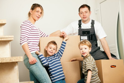 A family of four, including a woman in a striped long-sleeve top and two young children, are in a well-lit home environment with a staircase visible on the left. They are surrounded by several large cardboard boxes, some sealed and others partially open, filled with household belongings. The woman is holding one of the boxes while smiling at the camera, and the two young boys, dressed in striped shirts, are standing close by, one touching the box. A removal specialist from West Kensington Man with Van, wearing a white shirt and black protective overalls, stands behind the boxes, assisting with the moving process. The scene is set indoors, likely in a residential hallway or living area, with a neutral wall in the background and natural lighting emphasizing a professional yet personal atmosphere. This image reflects the process of house removals, packing, and family involvement in a relocation context within Kensington or nearby London areas, supporting local house moving and sustainable disposal of household items.