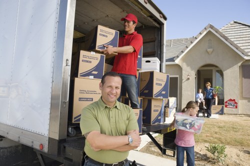 An outdoor scene showing two men involved in a house move on a residential street. Inside the back of a large white removals van, one man wearing a red cap and red t-shirt is standing on the van's loading area, handing a cardboard box to the other man, who is inside the van. The boxes are made of plain cardboard with printed labels, and appear to contain household belongings such as books or supplies. The man outside the van, dressed in a green shirt, is smiling with his arms crossed and standing in front of the vehicle, facing the camera. To the right, a woman in a pink top is holding a transparent plastic storage box with a purple lid, while another woman stands near a house with a white exterior, arched window, and tiled roof, observing the activity. The background shows a clear sky, a tidy lawn, and a row of similar houses, indicating a typical residential area possibly in West Kensington or nearby London postcode areas. The image reflects a professional house removal operation, emphasizing careful handling of household items during a local move in London.