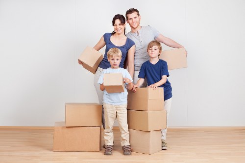 A family of four, including two young boys and their parents, standing in a spacious, well-lit room with a light-colored wooden floor and plain white wall, surrounded by several brown cardboard moving boxes of various sizes. The mother, dressed in a dark blue top, holds a small box, while the father, in a light grey t-shirt, stands behind her with his arm around her shoulder. The two boys, both wearing blue shirts, hold a box each at chest level, with one boy slightly in front and the other to his side. Additional boxes are stacked on the floor around them, some open and some closed, likely prepared for a house removal. The scene captures a moment of packing or unpacking, with a focus on the organized, clean environment typical of a UK residential move, possibly within the West Kensington or nearby London area serviced by West Kensington Man with Van. The image conveys the practicality and family-oriented nature of house removals, emphasizing careful handling of household items such as boxes and personal belongings in a domestic setting.