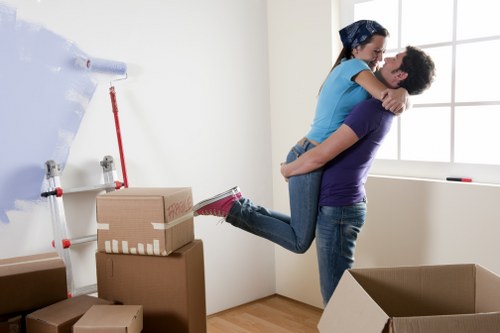 A young couple engaged in a joyful moment during a house move inside a well-lit room with white walls and a large window. The man, wearing a dark purple t-shirt and blue jeans, is standing on the floor, lifting the woman in a blue t-shirt and jeans. The woman has her arms around the man's shoulders and is smiling, appearing excited. In the background, there are several cardboard moving boxes of various sizes, some stacked on top of each other, with a few still sealed with packing tape. A roll of packing paper is mounted on the wall, along with a red-handled tool or stick leaning against it. The wooden flooring and simple white skirting boards are visible, indicating a residential setting in West Kensington. The scene captures a moment of packing and relocating within a typical house interior, emphasizing the involvement of a professional removals service by West Kensington Man with Van.