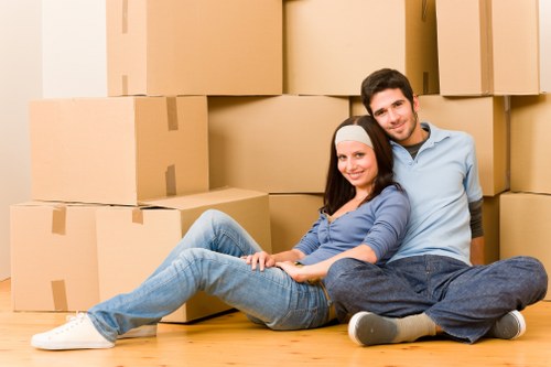 A young couple with light skin sitting on a light wooden floor inside a residential space, surrounded by numerous large cardboard packing boxes arranged in the background, some stacked to form a makeshift wall. The woman has long dark hair, wearing a white headband, a blue long-sleeved shirt, and light blue jeans, and is seated with her legs extended, smiling at the camera. The man has short dark hair, dressed in a light blue shirt and dark jeans, sitting with his legs bent and crossed at the ankles, also smiling. The setting appears to be a house or apartment ready for a move, with natural indoor lighting illuminating the scene. The presence of packed boxes indicates preparations for a house removal or relocation. The scene conveys a casual, relaxed atmosphere common during residential moves in West Kensington or nearby areas, and the image aligns with house removals services offered by West Kensington Man with Van.