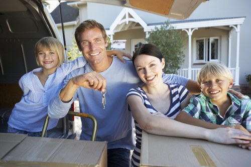 A family of four, comprising two adults and two children, are engaged in packing their belongings for a house move in front of a suburban home in West Kensington. The father, with light brown hair and a friendly smile, holds a set of keys in his right hand and is dressed in a light blue shirt. The mother, with short dark hair, wears a striped top and is sitting next to him, smiling at the camera. The two children, a girl with blonde hair on the father's left and a boy with blonde hair on the mother's right, are both smiling and appear excited about the move. All are standing behind an open van with its tailgate down, which contains neatly packed cardboard boxes and household items. Visible items include a cardboard box in the foreground on a wooden table or surface. The background shows a white, modern-style house with large windows and a small porch, surrounded by greenery. The scene is lit with natural daylight, suggesting a clear day, and reflects a typical residential relocation scene, consistent with West Kensington removals services.