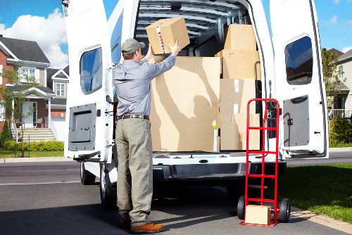 A professional removal specialist from West Kensington Man with Van, dressed in a grey shirt, beige trousers, and a cap, is seen unloading a cardboard box from the back of a white removals van parked on a residential street. The van's rear doors are open, revealing neatly stacked moving boxes of various sizes, some secured with packing tape. The boxes are made of brown corrugated cardboard and are arranged to allow easy access for unloading. A red hand truck with a black handle is positioned to the side of the van, with a single box resting on its platform, ready for transport. The background features modern suburban houses with well-maintained lawns, and the street appears clean and quiet under a partly cloudy sky. The scene captures elements typical of house removals in the West Kensington area, focusing on the careful handling of household belongings during a local move.