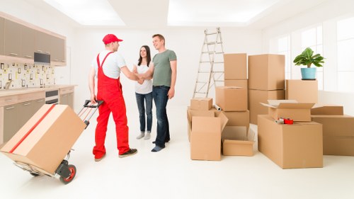 Inside a bright, modern residential room with white walls and large windows allowing natural light, a man dressed in red work overalls and a red cap is shaking hands with a smiling couple. The man in casual clothing, consisting of a grey T-shirt and dark jeans, is holding a box, while the woman, wearing a light-colored top and dark jeans, stands beside him with a friendly expression. Behind them, a tall aluminium step ladder leans against the wall, indicating recent or ongoing moving preparations. To the right, an assortment of packed cardboard boxes of various sizes are neatly stacked on the floor, some labeled, along with a small red tape measure placed atop one of the boxes. An indoor potted plant with green foliage sits on a windowsill, adding a touch of decor to the space. Additionally, visible kitchen cabinetry in the background features a contemporary design with neutral tones, completing the scene of a house being prepared for a move, consistent with services offered by West Kensington Man with Van, based near Kensington in London, as implied by the setting and context of the removals process.
