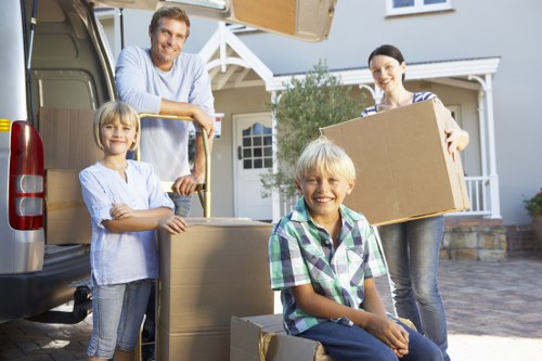 A family of four with light skin are engaged in a house move outside a suburban home. The father, wearing a light blue shirt, stands in the back near an open grey removals van, smiling as he looks toward the camera. To his right, the mother, dressed in a striped top, holds a large cardboard box, also smiling. In front, a young boy with blonde hair, wearing a green and white checkered shirt, is sitting on a cardboard box on the driveway, smiling at the camera. Next to him, a girl with blonde hair, dressed in a light-colored shirt, stands with her arms crossed, smiling. The driveway is paved, and the background features a modern white house with a porch, a gabled roof, and some greenery. The scene is brightly lit, suggesting daylight, with the family actively involved in packing and loading household items, typical of a professional removals service in the west London area.