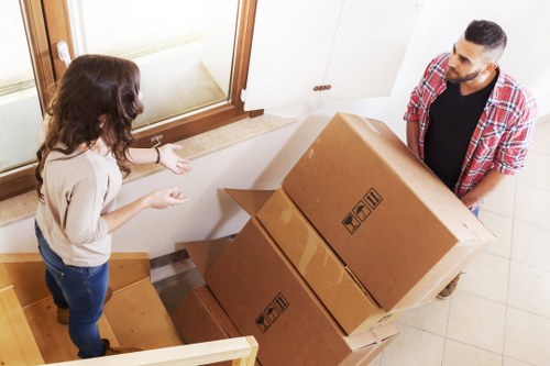 A male moving professional from West Kensington Man with Van transfers three large cardboard boxes in a residential property, located at the bottom of a staircase near an entrance door with a large window. The boxes are brown with black handling symbols and are stacked, with the top box slightly tilted. The man is wearing a red and white checkered shirt over a black T-shirt, and stands on a tiled floor, supporting the boxes as he prepares to carry them. A woman with long, wavy brown hair, dressed in a beige sweater and blue jeans, stands nearby on the staircase, engaging in conversation with the mover. The interior features a wooden staircase, white walls, and a cream-colored door frame, indicating a typical UK house interior in an area like West Kensington or nearby. The natural light from the window illuminates the scene, emphasizing the textures of the cardboard, clothing, and wood surfaces, reflecting a professional and organized house removal process.