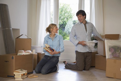 In a bright, airy room with large windows allowing natural light and a view of greenery outside, a woman and a man are engaged in packing or unpacking during a house move. The woman, with blonde hair tied back and wearing a light blue shirt and dark jeans, is kneeling on the wooden floor and holding a stack of books. The man, with short dark hair and dressed in a striped button-up shirt and khaki trousers, is kneeling beside open cardboard boxes, appearing to be preparing or organizing belongings. The room contains several packed moving boxes, some filled with household items such as books and soft furnishings, and others partially open, revealing contents like fabric and plastic wrapping. The scene captures a domestic setting typical of a residential house in West Kensington, with the focus on the furniture, cardboard boxes, and personal items involved in a house removal or relocation process, reflecting the services of West Kensington Man with Van in a professional and factual manner.