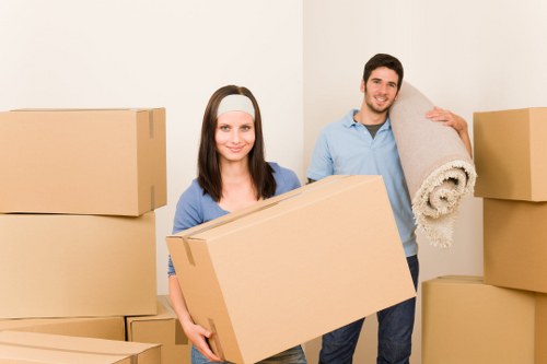 A young woman and a young man are standing in a clean, well-lit room with white walls, surrounded by several large cardboard boxes, indicating a house move. The woman has shoulder-length dark hair, is wearing a light blue blouse and a light grey headband, and is smiling while holding a medium-sized cardboard box in front of her. The man next to her has short dark hair, is dressed in a light blue shirt and dark trousers, and is smiling as he carries a rolled-up light-colored rug over his shoulder. The boxes vary in size, some stacked to the sides, and they occupy most of the foreground and background, suggesting an active packing or moving process typical of house removals in West Kensington or nearby areas. The setting appears to be an interior residential space, with minimal background details, emphasizing the activity of packing items for a household relocation part of a professional removals service.
