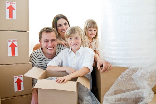 A family of four, consisting of a man, woman, and two young children, inside a bright, indoor residential space surrounded by stacked cardboard moving boxes with red and white directional arrows, indicating careful handling and packing for a home move. The family members are smiling and appear to be in a positive mood, with the young boy sitting in an open cardboard box in the foreground, and the other family members standing behind him, slightly leaning forward. The boxes are made of brown corrugated cardboard and some are partially filled with packing material such as bubble wrap. To the right, there is a larger box wrapped in protective packaging, potentially for furniture or fragile items, suggesting a household move within a residential area, possibly in West Kensington or nearby in London. The environment has natural lighting, indicating daylight coming through a window or doorway, and the background includes a white wall and a door or curtain, creating a clean, organized scene typical of home removals and relocation preparations as offered by West Kensington Man with Van.
