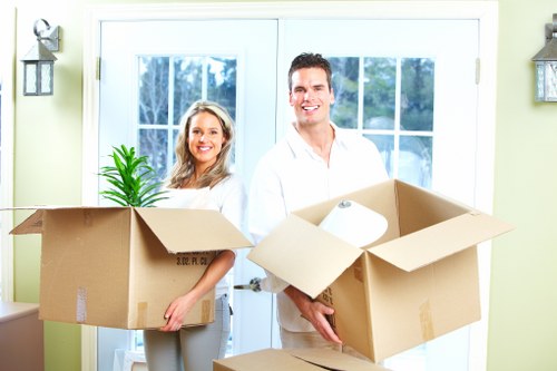 A man and woman stand inside a residential home during a house removal, holding large open cardboard boxes filled with packing materials and household items. The woman, positioned to the left, has shoulder-length blonde hair and is smiling while holding a box with a small potted plant visible inside. The man, on the right, has short dark hair and is also smiling as he holds a box containing rolled-up paper or fabric. Behind them, a set of white-framed glass doors provides natural light, revealing glimpses of greenery outside. The light green walls are decorated with two wall-mounted lantern-style light fixtures on either side of the doors. The scene takes place in a bright, tidy interior, indicating a professional house removal carried out by West Kensington Man with Van, serving the Kensington area. The image showcases the careful packing and handling typical of a domestic moving service, with attention to household furniture, boxes, and packing essentials, emphasizing the company's expertise in residential relocations within West Kensington and nearby localities.