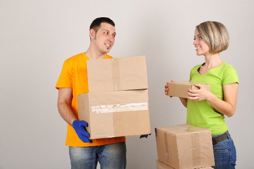 A professional moving scene featuring a male and female mover inside a residential property, possibly in West Kensington or nearby. The male mover, dressed in a bright yellow T-shirt and wearing blue protective gloves, is holding a large cardboard box with clear tape securing the flaps, and looks engaged in conversation with the female mover. The female, wearing a lime green T-shirt and blue jeans, is smiling as she holds a smaller cardboard box, standing next to a stack of other packed boxes on the floor. The background is a plain, light grey interior wall, indicating a clean, neutrally decorated room. Both movers are positioned on a smooth, light-colored floor, and natural lighting illuminates the scene, highlighting the clarity and texture of the cardboard boxes and their contents. The scene underscores professional house removal services, with attention to careful handling of household items such as boxes and personal belongings, suitable for a service offered by West Kensington Man with Van in the West Kensington area, part of their removals service portfolio.