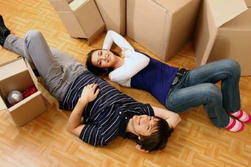 A young couple lies on their living room floor amidst open cardboard boxes, surrounded by packed and unpacked household items. The woman, wearing a navy and white long-sleeve shirt and jeans, is lying on her back with her head slightly tilted and eyes closed, with her hands resting behind her head. The man, dressed in a striped polo shirt and grey trousers, is lying on his side, facing the ceiling, with one arm resting on his chest and the other on his hip. The floor has a wood-effect finish, and several boxes are visible around them, some closed and others open, showing household items like a red and a silver ball. The scene suggests a house relocation within a residential street, possibly in West Kensington or nearby London areas, with the presence of a removals van visible outside through a window or doorway, typical of a professional house removal service. The natural lighting and relaxed posture of the couple reflect the process of packing or unpacking during a house move, with the focus on household packing essentials and furniture to be transported.