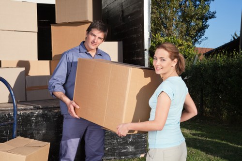 A male and female mover from West Kensington Man with Van are outdoors near a residential property, loading or unloading a large cardboard box. The man, dressed in a light blue shirt and dark trousers, is holding the box on one side, while the woman, in a light blue top and beige trousers, supports the other side. Behind them, an open black van filled with stacked cardboard boxes of various sizes is parked on a paved driveway. The boxes appear to be made of brown corrugated cardboard, some slightly crushed or taped, ready for moving. The scene is set in a well-maintained garden area with green grass, trees, and residential houses visible in the background under clear daylight conditions. The image conveys a professional, organized house removal operation in a suburban UK setting, subtly emphasizing local service expertise in residential relocations and removals.