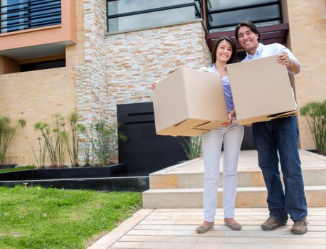 A young couple standing outdoors in front of a modern residential building during daylight hours, each holding a large cardboard moving box. The woman, with long dark hair, is wearing a light purple blouse and light grey trousers, while the man, with short dark hair, is dressed in a white shirt and dark jeans. They are smiling and appear to be in the process of moving into or out of a property in a suburban area, suggesting a house removals scenario in West Kensington. The background features a multi-story building with beige brick and stone exterior finishes, balconies with glass railings, and a small, landscaped garden with green grass, a few small plants, and paved pathway leading to the front steps of the house. The scene is well-lit with natural daylight, displaying a clean, organized environment suitable for domestic relocation services by West Kensington Man with Van.