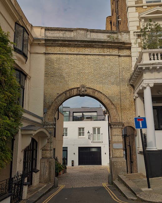 A narrow street in Earl's Court with an architectural archway made of beige brickwork, visible at the top of a small cobbled road. On the left side, there is a building with a white facade and curved 