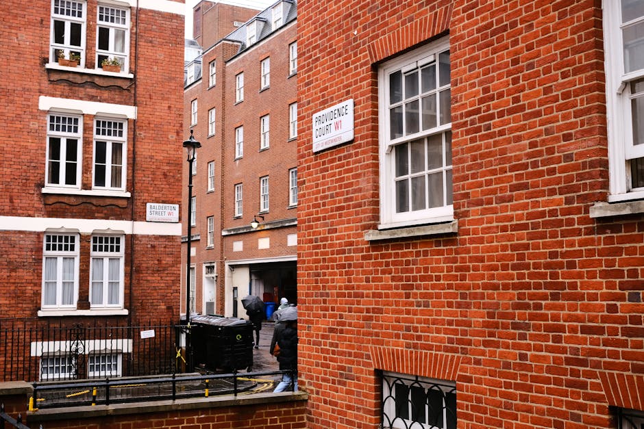 A narrow street in Earl's Court with an architectural archway made of beige brickwork, visible at the top of a small cobbled road. On the left side, there is a building with a white facade and curved 