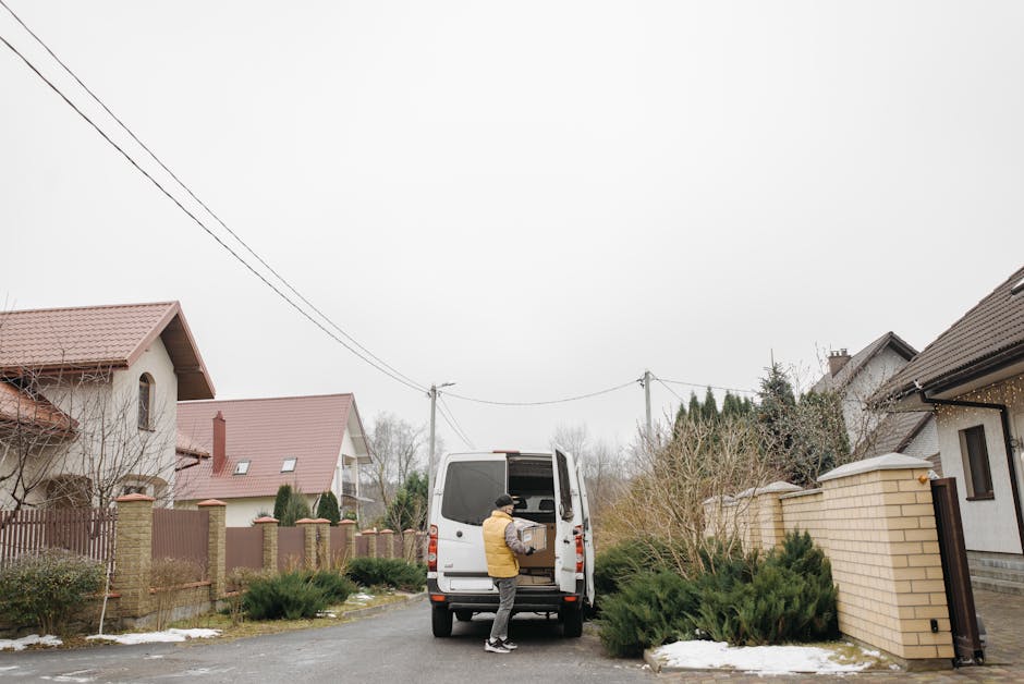 A person wearing a yellow jacket and black hat loading a cardboard box into the back of a white van parked on a suburban street, surrounded by residential houses with pitched roofs and brick walls, so