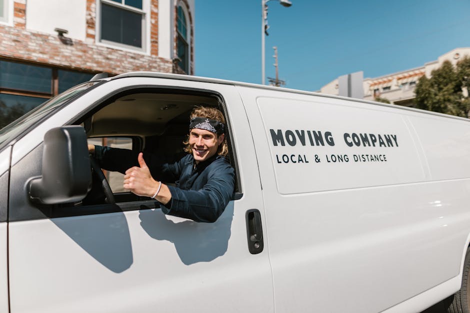 A person wearing a yellow jacket and black hat loading a cardboard box into the back of a white van parked on a suburban street, surrounded by residential houses with pitched roofs and brick walls, so